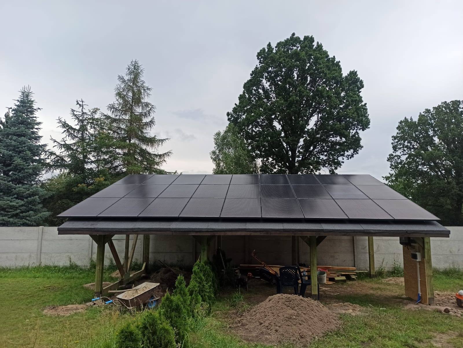 Zadaszenie z panelami fotowoltaicznymi w ogrodzie, widok na moduły w czarnych ramach, betonowe ogrodzenie w tle, otoczenie z drzewami i elementami budowlanymi.