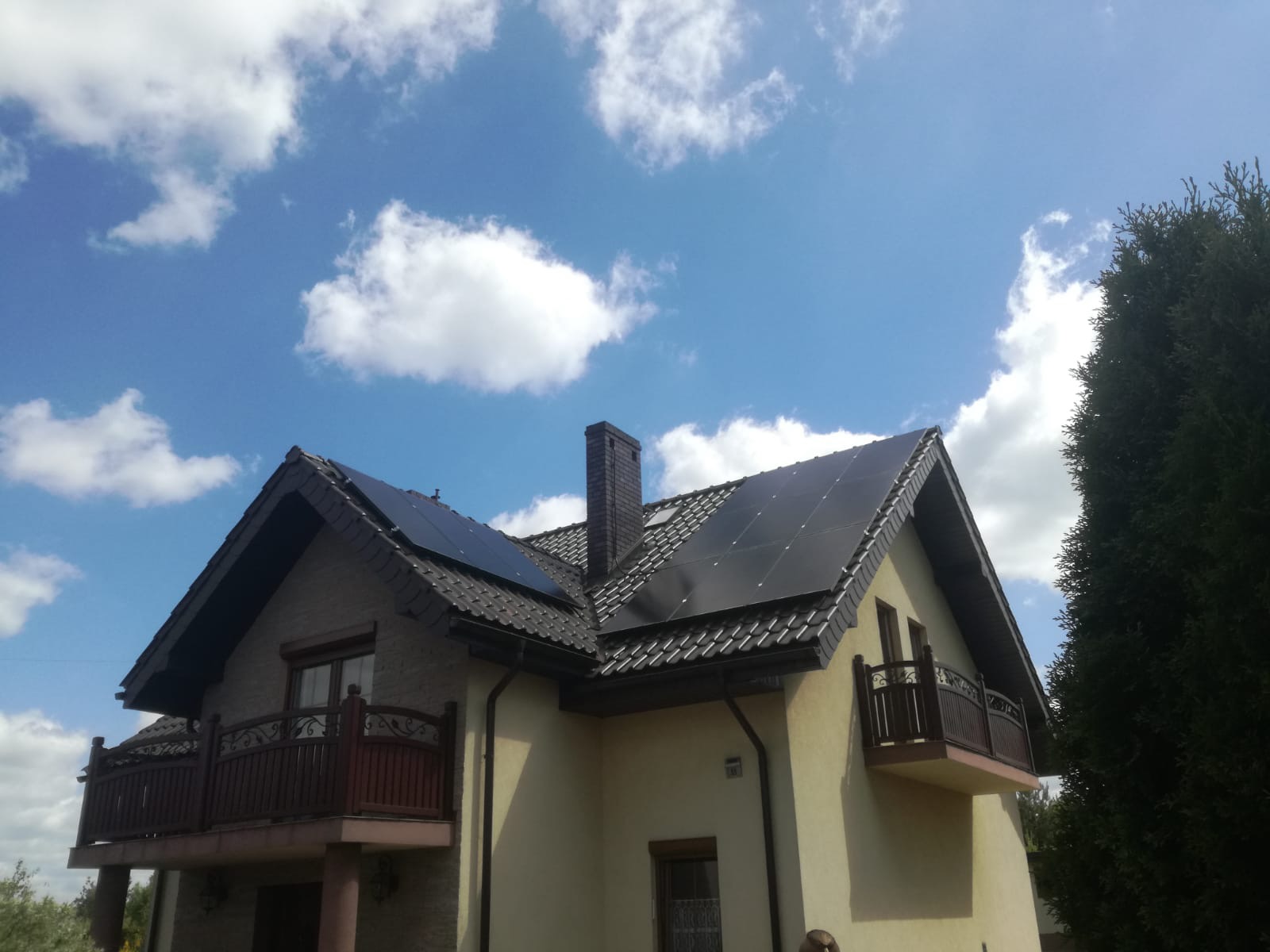 Panele słoneczne na dachu dwupiętrowego domu z balkonami, widok z dołu na tle błękitnego nieba z białymi chmurami.