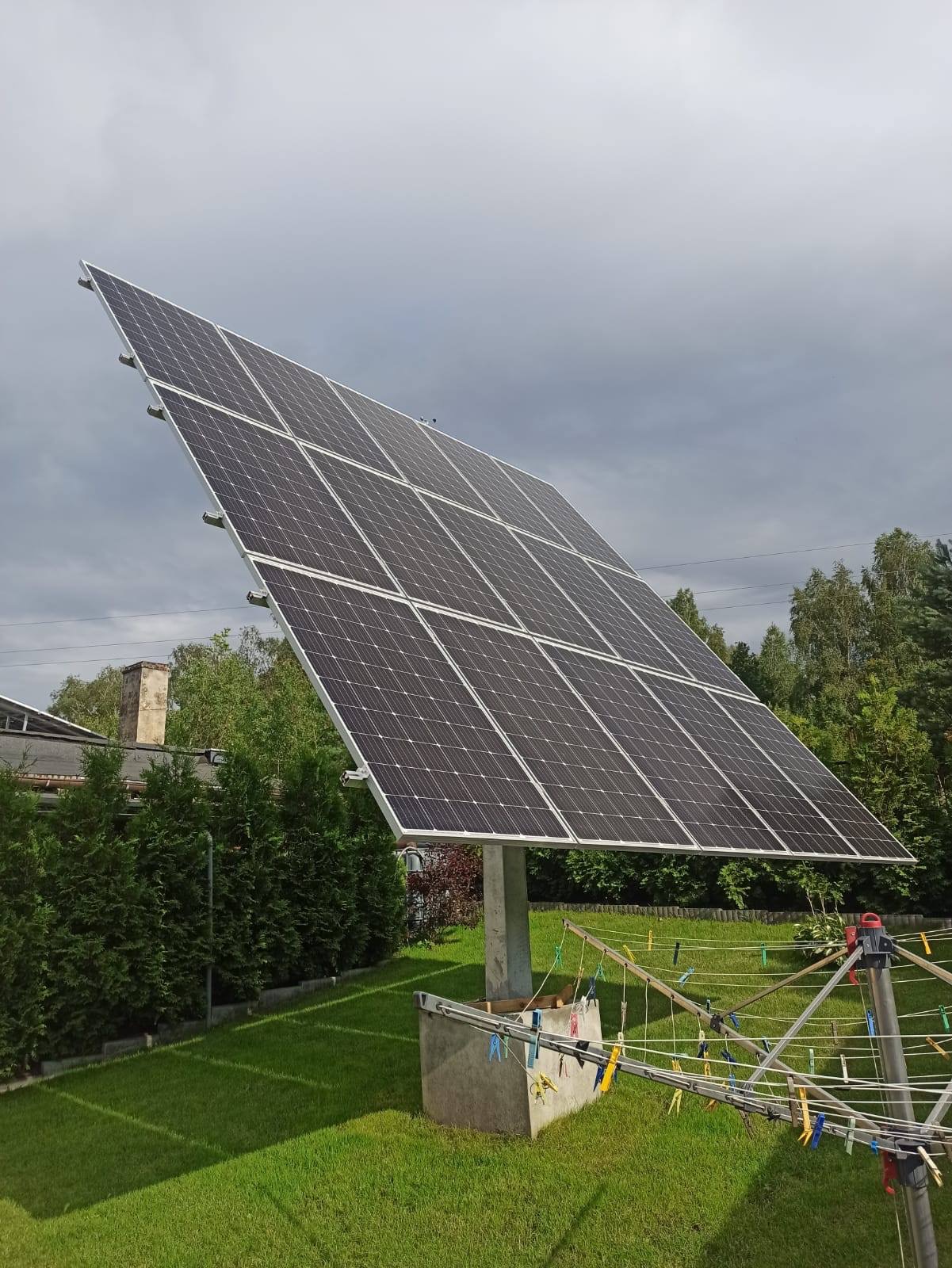 Wolnostojąca instalacja paneli słonecznych na betonowym fundamencie, widoczna na tle zielonego trawnika i pochmurnego nieba, z suszarką na pranie w pierwszym planie.