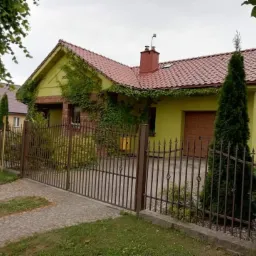 Żółty dom z czerwoną dachówką, częściowo porośnięty bluszczem, widziany zza metalowego ogrodzenia.