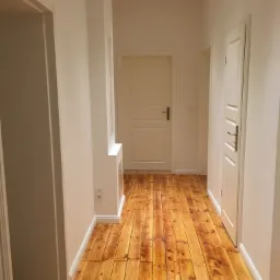 Jasny korytarz z odnowioną, lakierowaną podłogą z desek w odcieniach miodu i bursztynu, białe ściany z listwami przypodłogowymi, trzy białe drzwi z mosiężnymi klamkami.