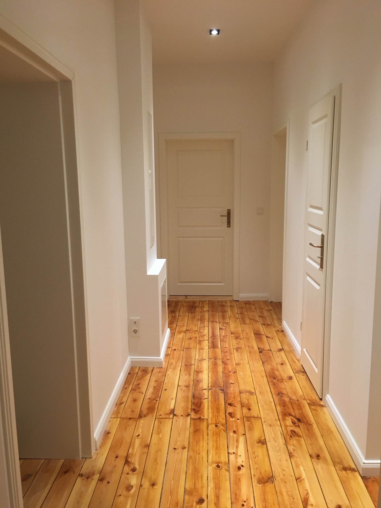 Jasny korytarz z odnowioną, lakierowaną podłogą z desek w odcieniach miodu i bursztynu, białe ściany z listwami przypodłogowymi, trzy białe drzwi z mosiężnymi klamkami.