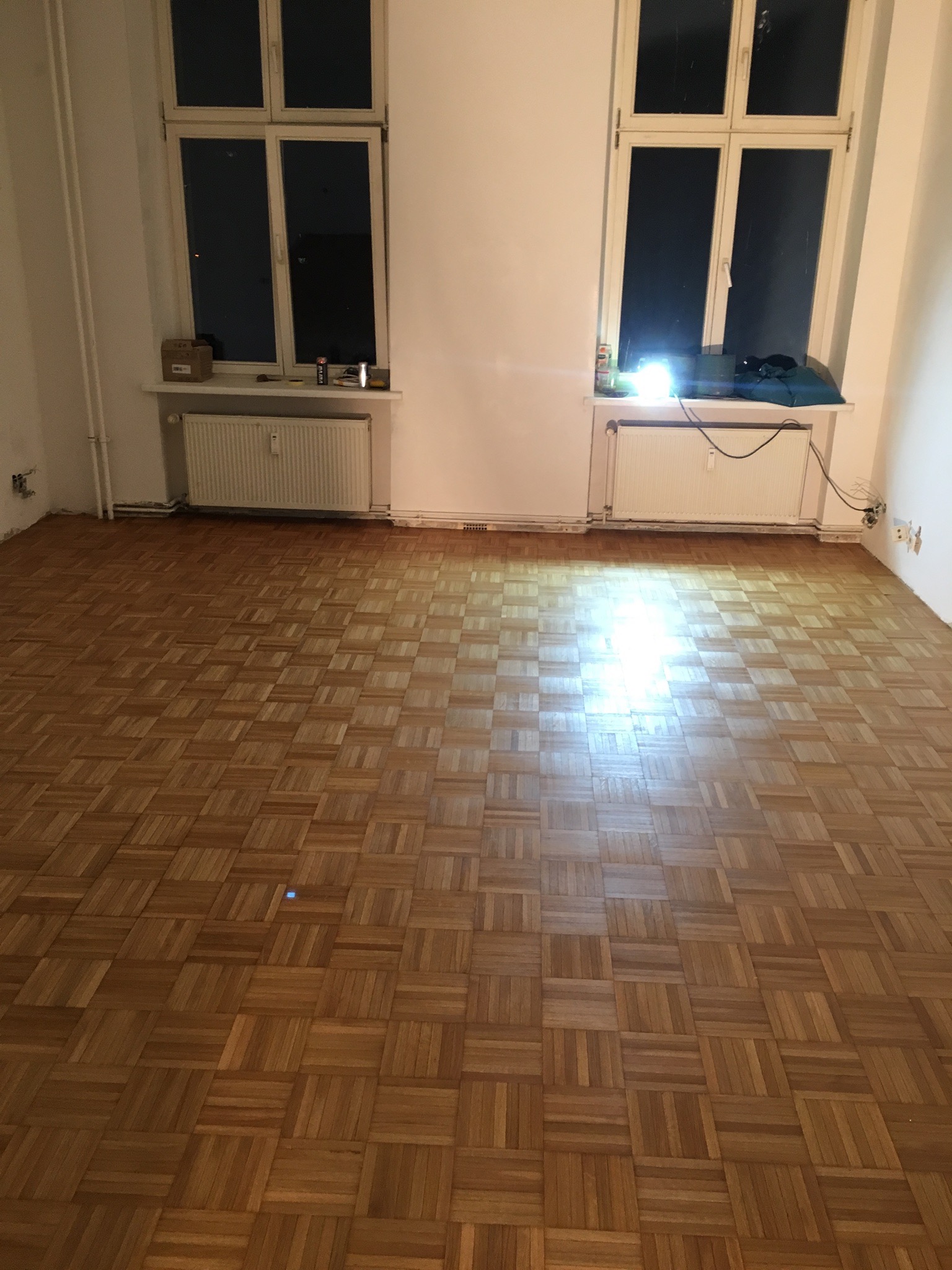Parkiet ułożony w kwadraty, widok na całe pomieszczenie z dwoma oknami, kaloryferami i białymi ścianami.