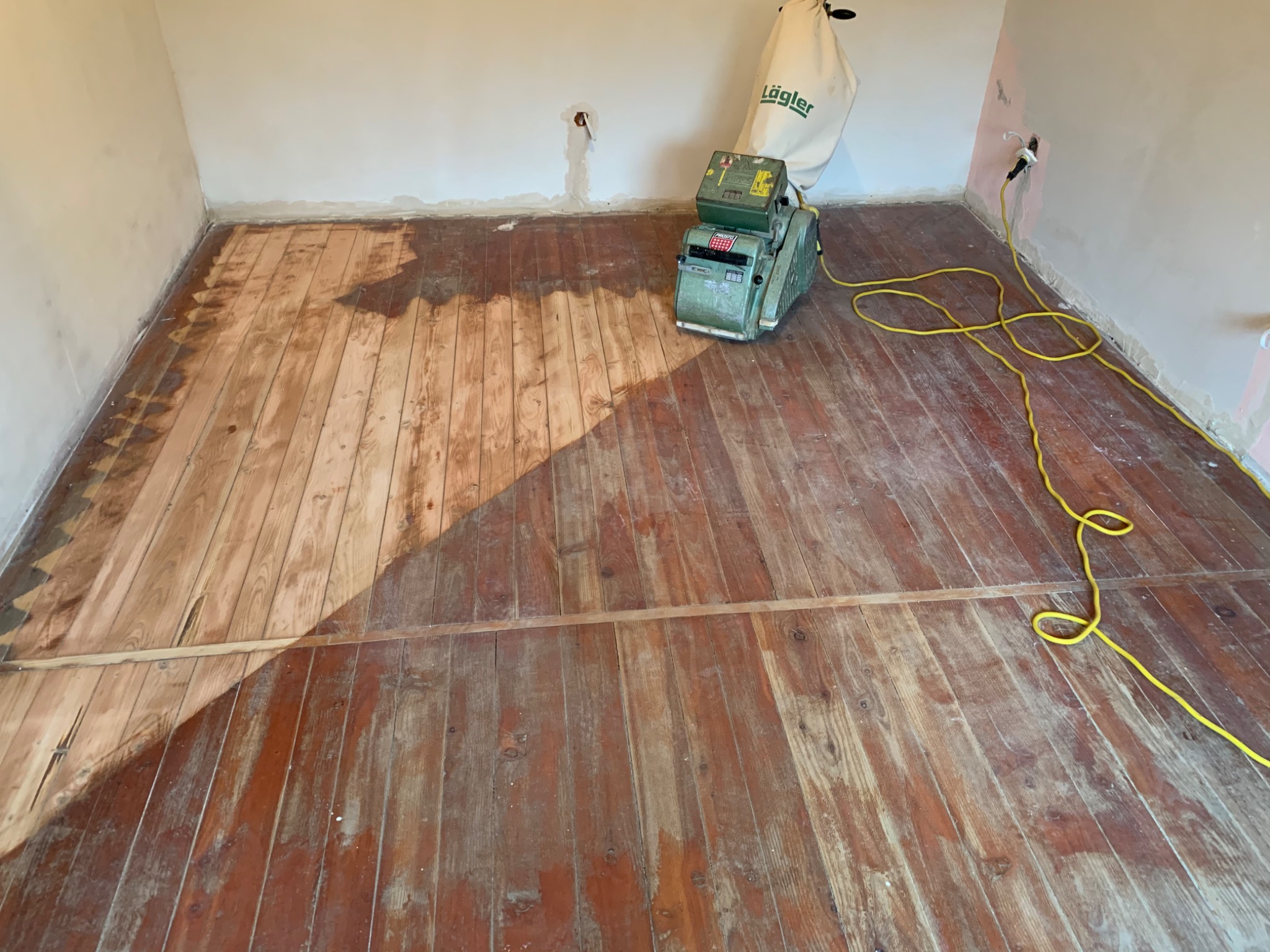 Fragment drewnianej podłogi w trakcie cyklinowania; widoczna maszyna do cyklinowania marki Läglera i obszar podłogi po obróbce, kontrastujący z surowym, ciemniejszym drewnem.