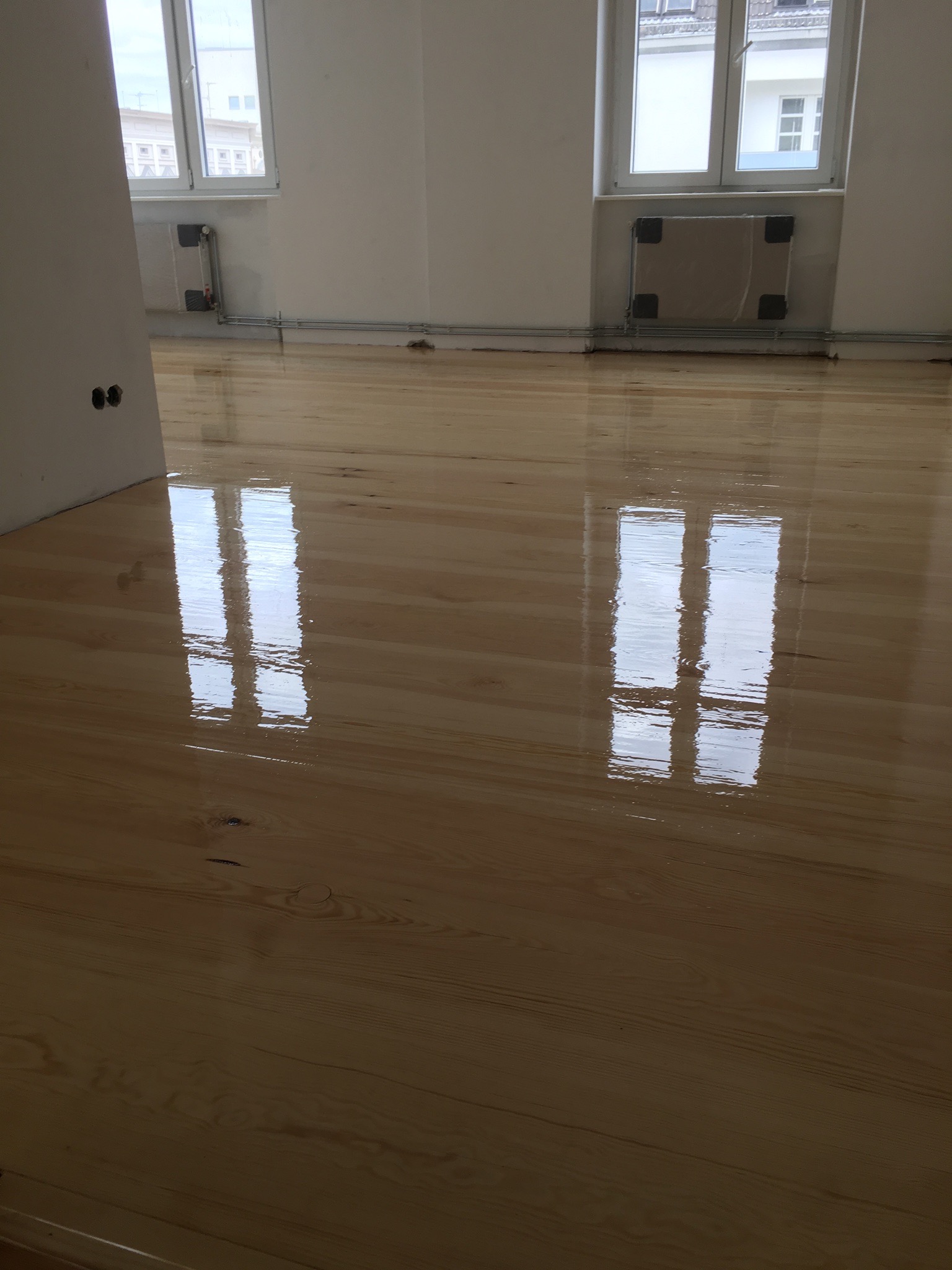 Błyszcząca, świeżo polakierowana podłoga parkietowa w jasnym odcieniu drewna, z odbiciami okien i białych ścian w pustym pokoju.