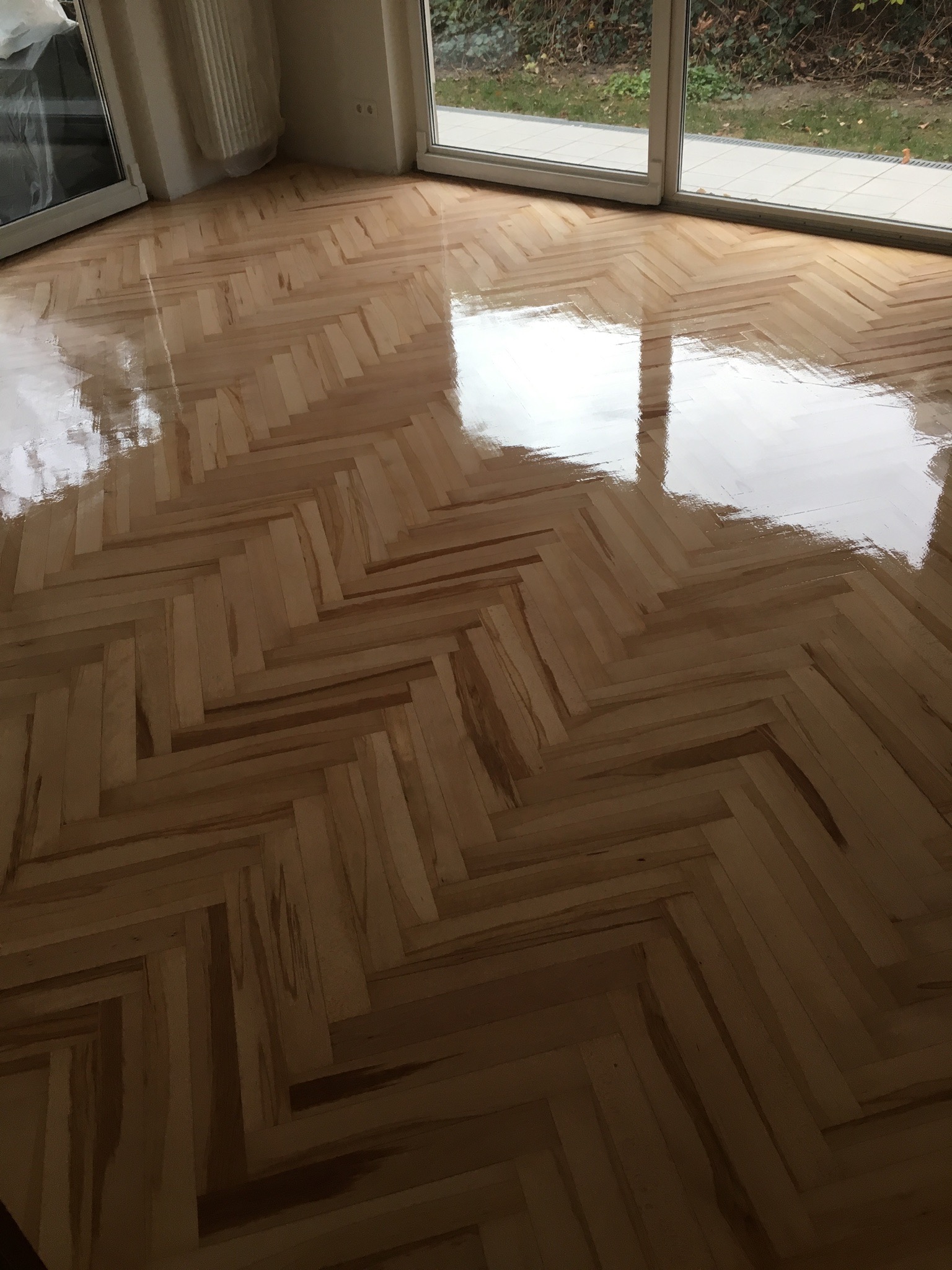 Świeżo ułożony, lakierowany parkiet w jodełkę, odbijający światło okien balkonowych w pustym pokoju.