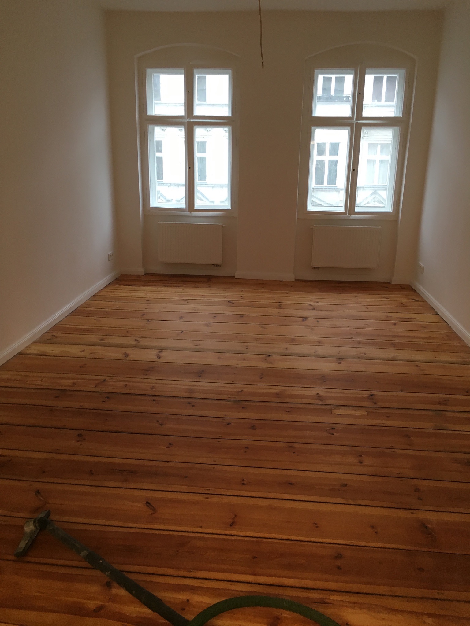 Wycyklinowana podłoga z desek w pustym pokoju z dwoma oknami, widoczny fragment szlifierki do parkietu.