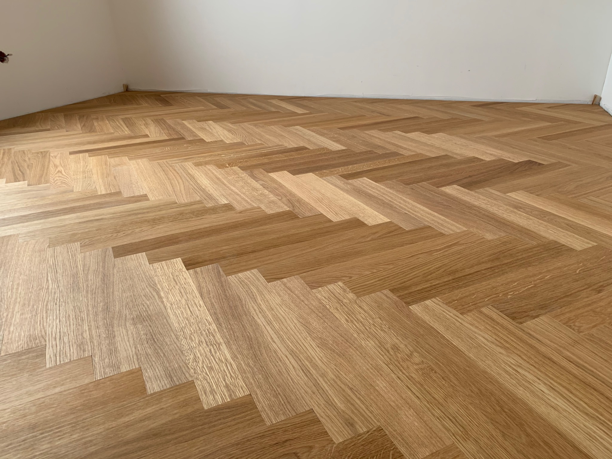 Ułożony parkiet w jodełkę francuską z widoczną strukturą drewna, przy białej ścianie z niedokończonymi listwami przypodłogowymi.