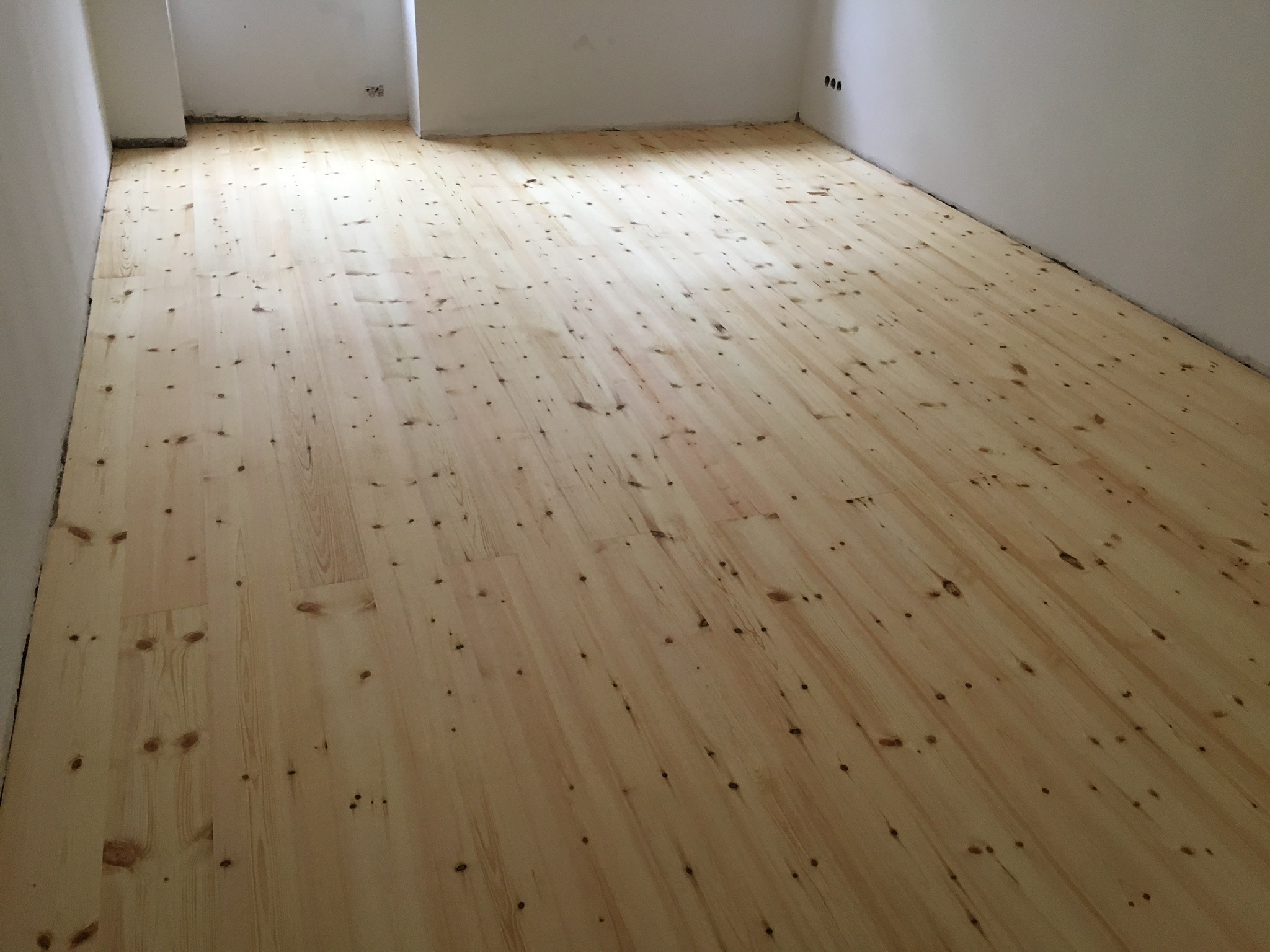 Jasne, sosnowe deski parkietowe ułożone w nowym pomieszczeniu z białymi ścianami i widocznymi gniazdkami elektrycznymi.