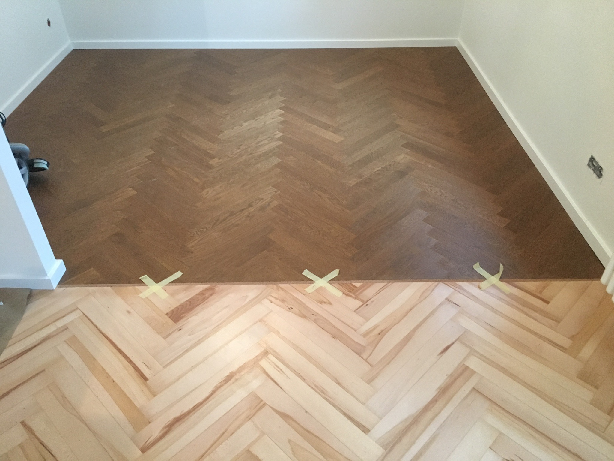 Fragment podłogi w trakcie układania parkietu w jodełkę, widoczna część już polakierowana i surowa, zabezpieczona taśmą malarską, białe listwy przypodłogowe
