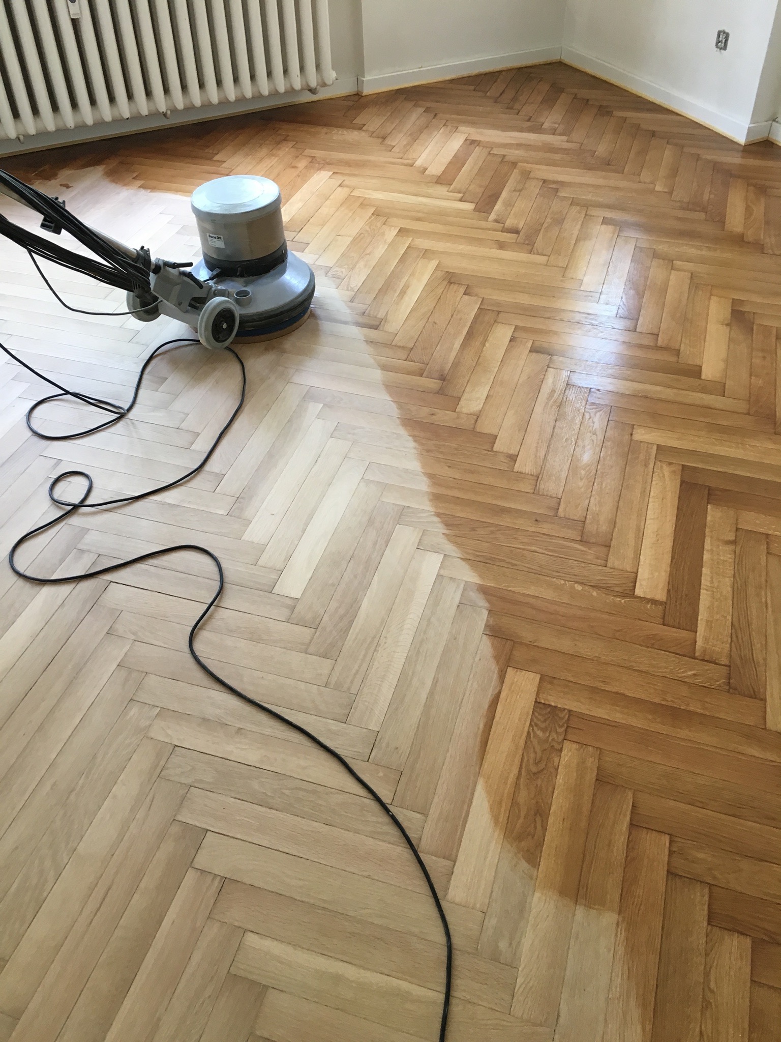 Szlifowanie parkietu jodełka maszyną, widoczna różnica w kolorze parkietu przed i po szlifowaniu, kabel maszyny na podłodze.
