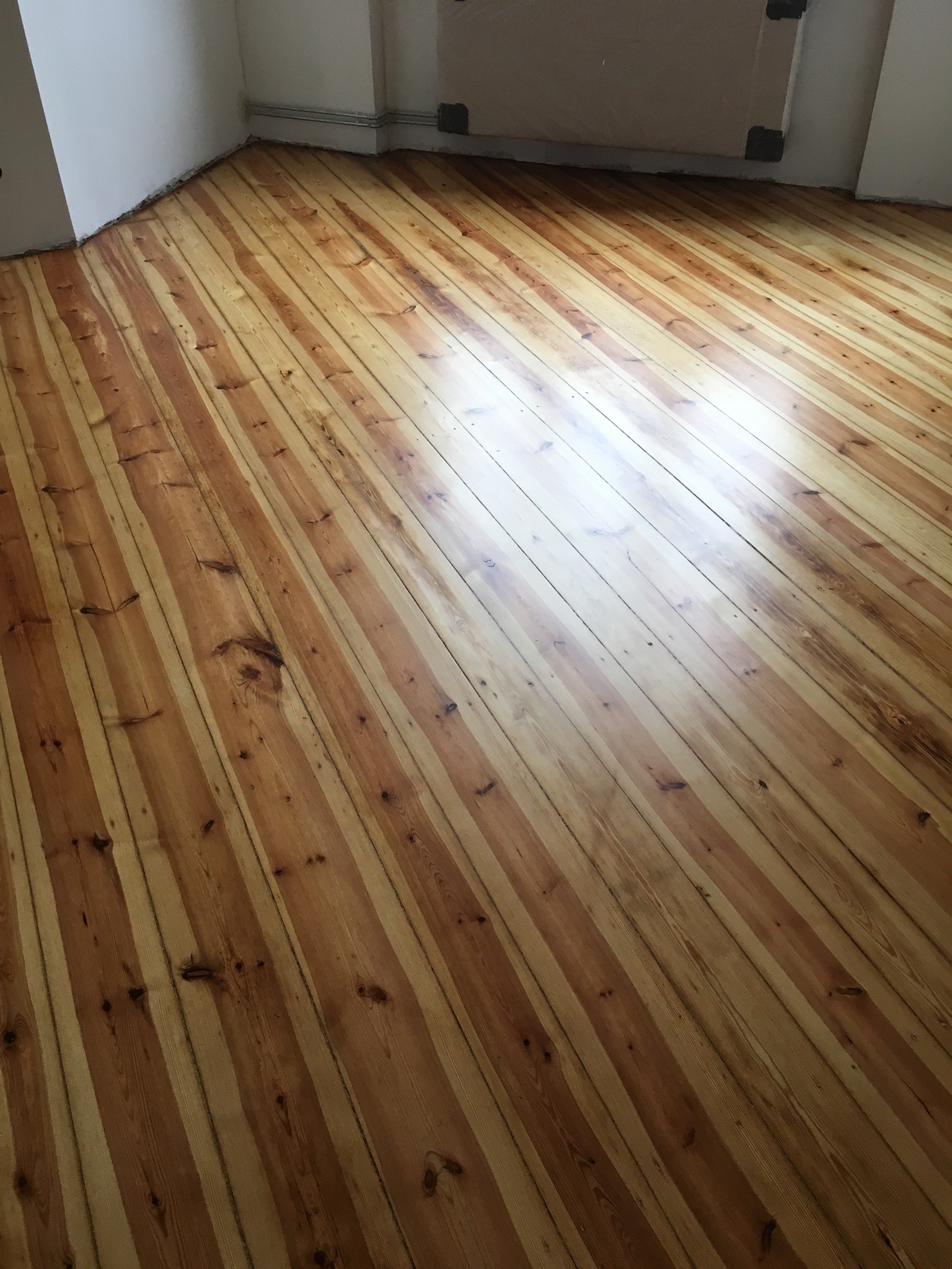 Wykończona podłoga z jasnego drewna z widocznymi słojami, ułożona ukośnie w jasnym pomieszczeniu z białymi ścianami i listwami przypodłogowymi.