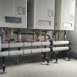 Trzy białe kotły grzewcze marki Immergas zamontowane na ścianie z białych bloczków, z widoczną instalacją rur i zaworów, oraz zmiękczacz wody w folii po lewej stronie.