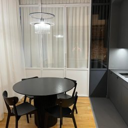 MONSTER - WELD MICHAŁ BERNATOWICZ - Nowoczesna jadalnia: czarny okrągły stół z krzesłami na drewnianej podłodze, przeszklona ściana z białymi ramami i zasłonami, designerska lampa wisząca.
