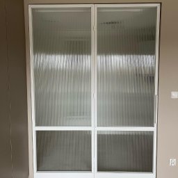 MONSTER - WELD MICHAŁ BERNATOWICZ - Nowoczesne, przesuwne drzwi z ryflowanego szkła w białej ramie, widoczne na tle parkietu w jodełkę i ściany w odcieniach beżu. Minimalistyczny design.