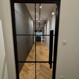 MONSTER - WELD MICHAŁ BERNATOWICZ - Nowoczesne, czarne, metalowe drzwi loftowe z szybami, widok na parkiet ułożony w jodełkę. Minimalistyczne wnętrze z szarą ścianą i widocznym oświetleniem.