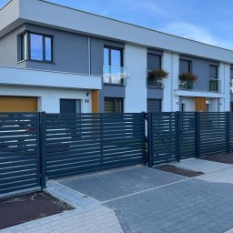 MONSTER - WELD MICHAŁ BERNATOWICZ - Nowoczesne, poziome ogrodzenie metalowe w kolorze grafitowym przed szeregową zabudową. Widoczne skrzydło bramy i furtka. Kostka brukowa na podjeździe.
