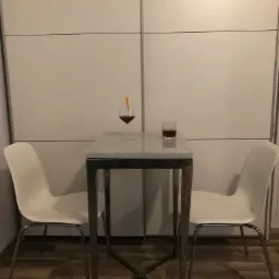 Stół barowy z marmurowym blatem i metalową konstrukcją, otoczony dwoma białymi krzesłami. Na blacie stoją dwa drinki: jeden w kieliszku z dekoracją z pomarańczy, drugi w szklance typu old fashioned...