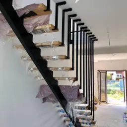 Metalowe schody z drewnianymi stopniami w trakcie montażu, zabezpieczone folią, z czarną, ażurową balustradą i widocznym fragmentem okna balkonowego w tle.