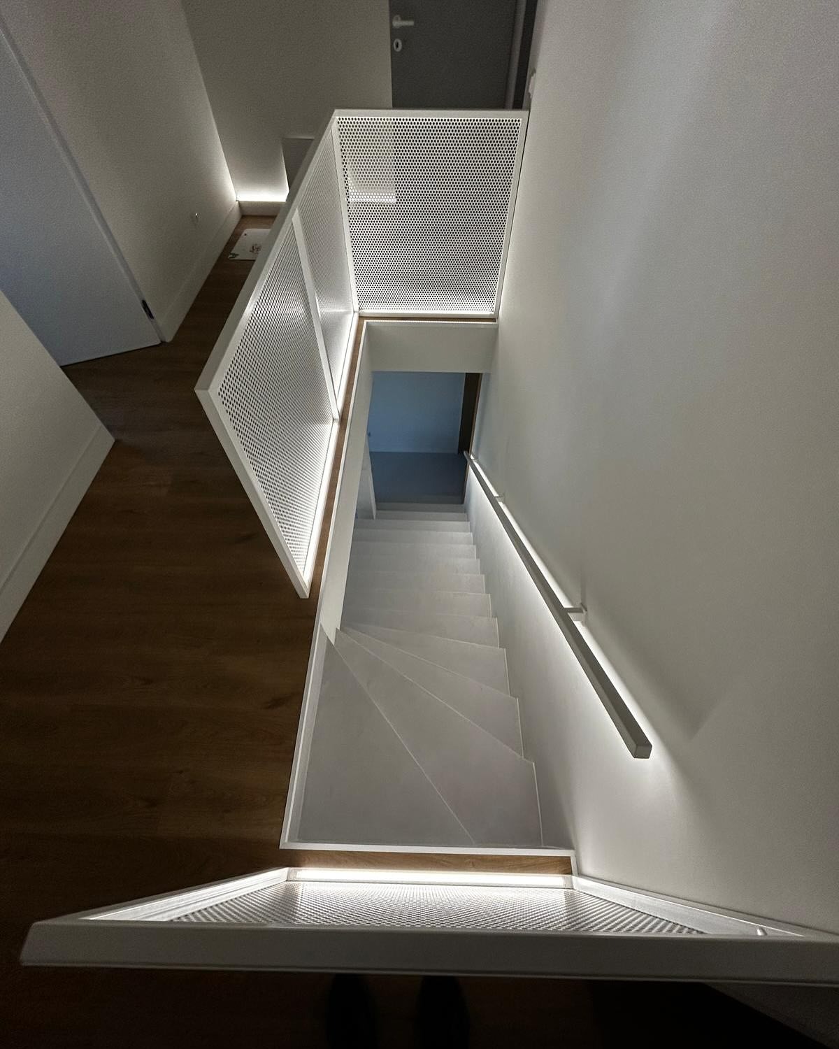 Nowoczesne, białe schody metalowe z perforowaną balustradą i subtelnym oświetleniem LED, widziane z góry. Minimalistyczny design, drewniana podłoga w tle.