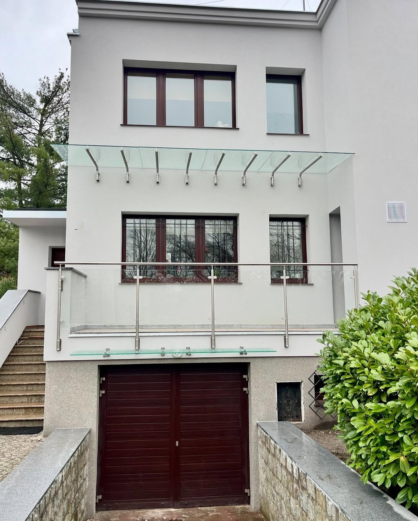 Nowoczesny dom z przeszkloną balustradą balkonową i zadaszeniem nad wejściem do garażu. Elementy metalowe w kolorze srebrnym, brązowe okna i brama garażowa.