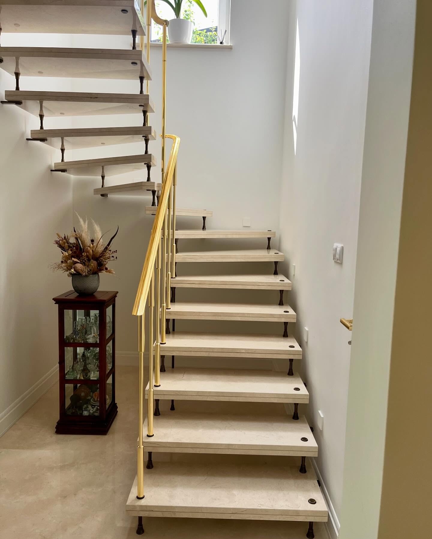 Nowoczesne schody z jasnego drewna i złotą balustradą, minimalistyczny design, widok z dołu, dekoracyjny bukiet w tle, subtelne oświetlenie.