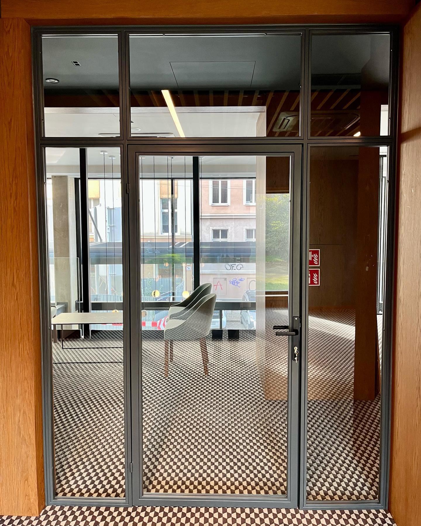 Szklane drzwi z metalową ramą, widok na wnętrze biura z krzesłami i wzorzystą podłogą. Nowoczesny design, naturalne światło, detale wykończenia.