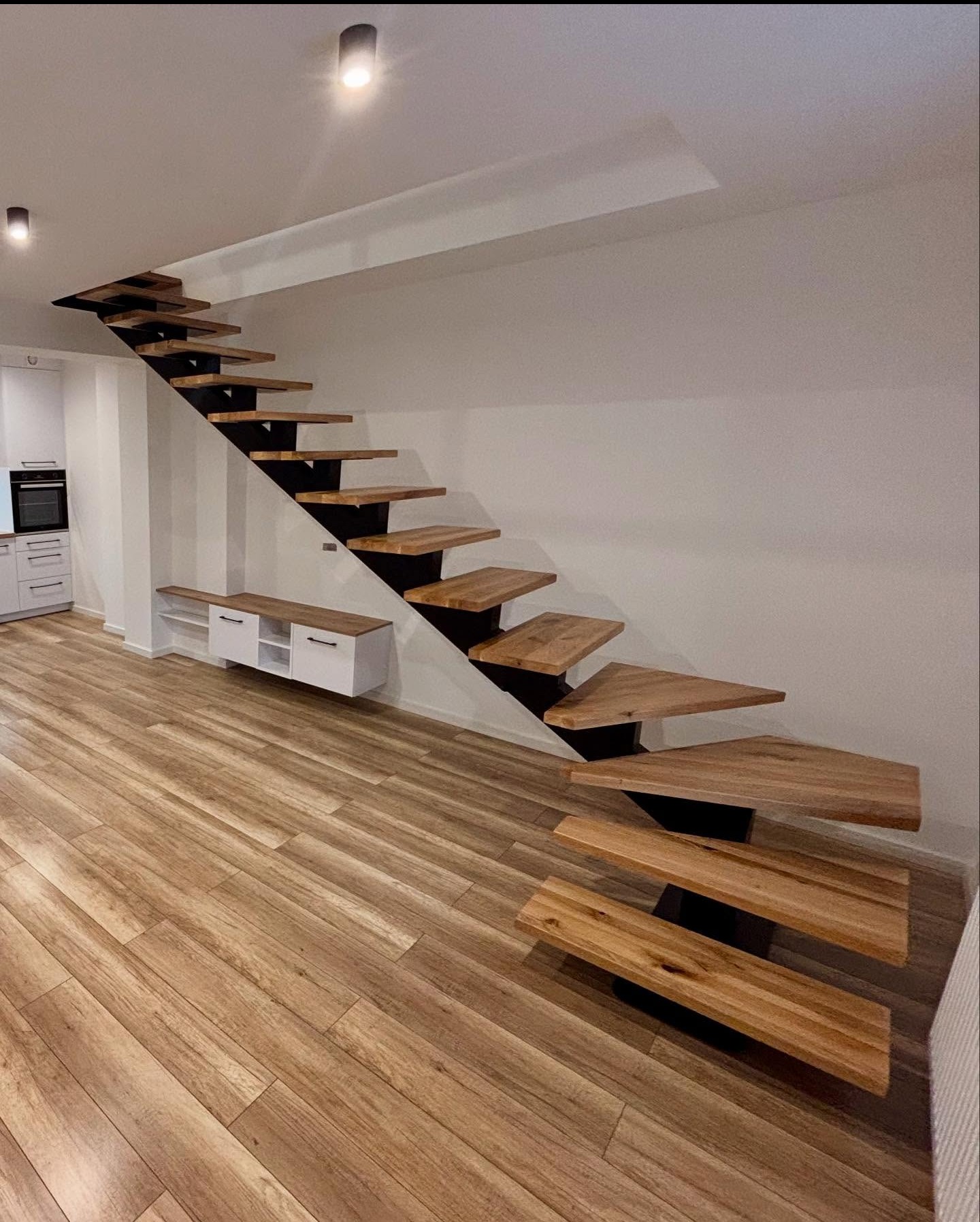 Nowoczesne schody z drewnianymi stopniami i czarną metalową konstrukcją, zamontowane w jasnym wnętrzu z drewnianą podłogą i białymi meblami.