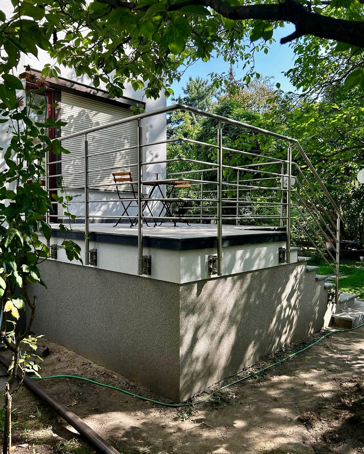 Nowoczesny taras z metalową balustradą ze stali nierdzewnej, widok z dołu, zieleń w tle. Minimalistyczny design, szara elewacja budynku, elementy małej architektury.