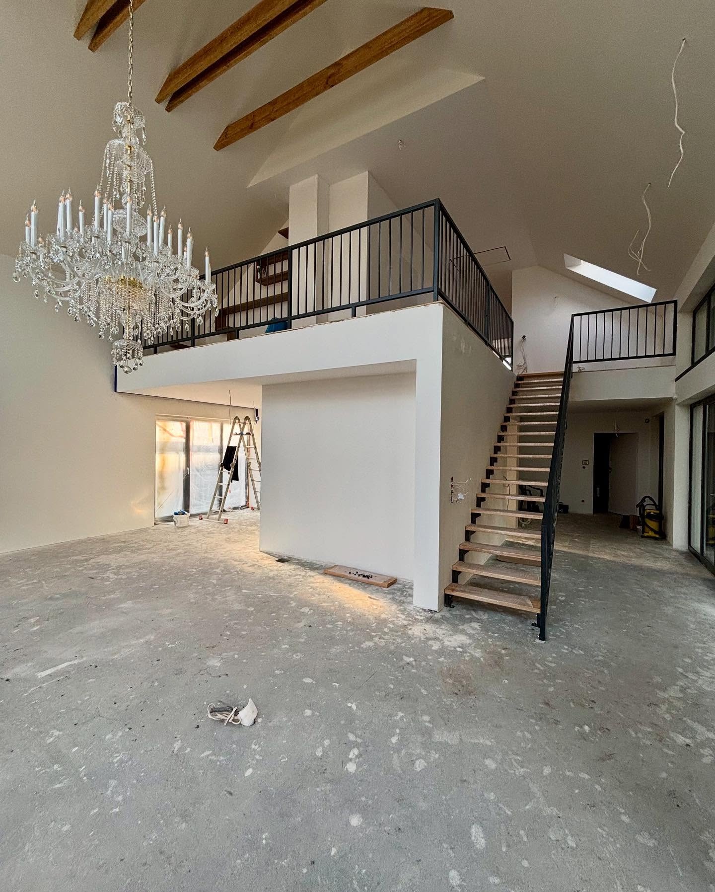 Wnętrze domu w trakcie wykańczania: drewniane schody z czarną balustradą, antresola z barierką, żyrandol kryształowy, surowe ściany i podłoga, drabina oparta o okno.