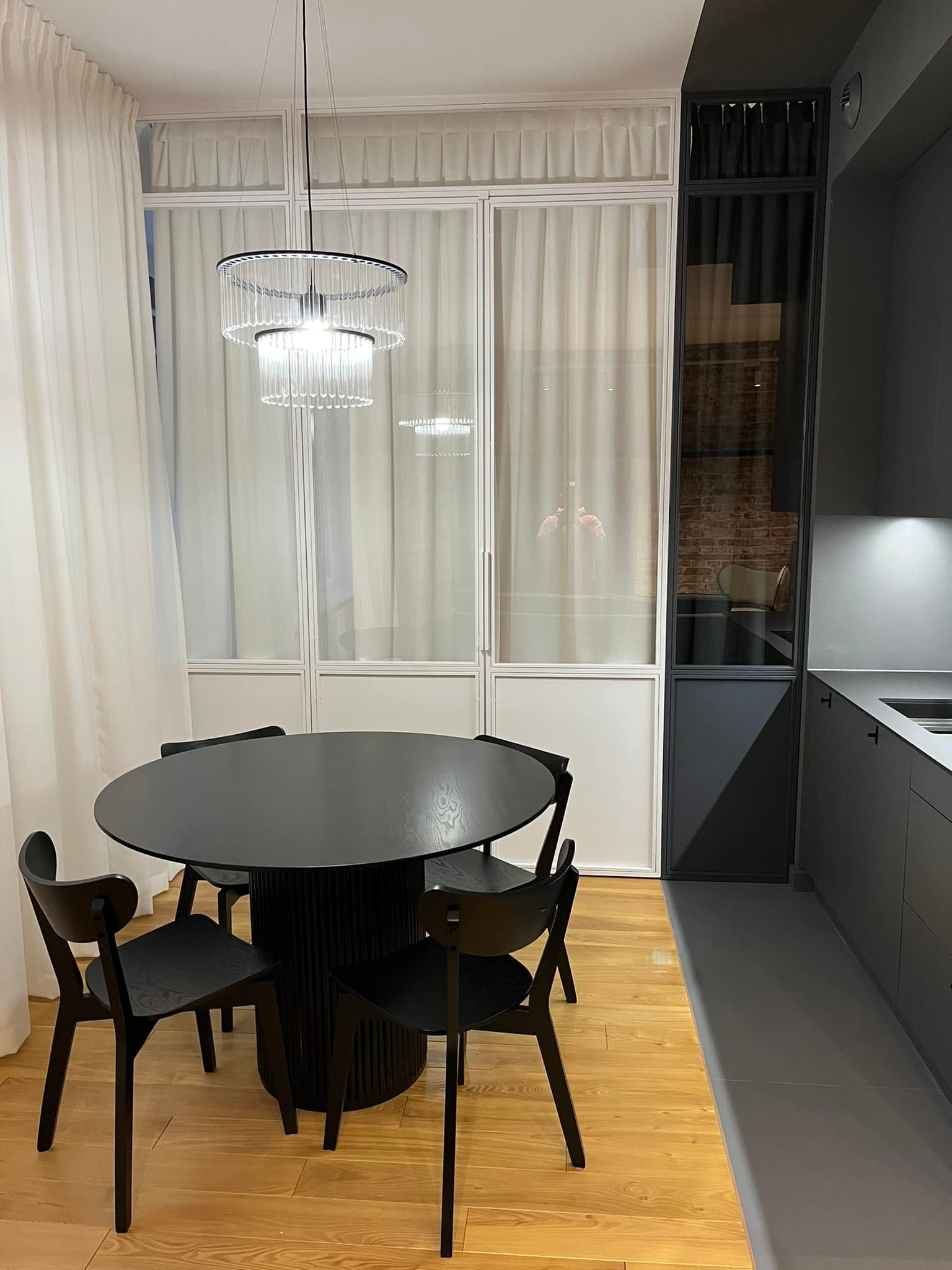 Nowoczesna jadalnia: czarny okrągły stół z krzesłami na drewnianej podłodze, przeszklona ściana z białymi ramami i zasłonami, designerska lampa wisząca.