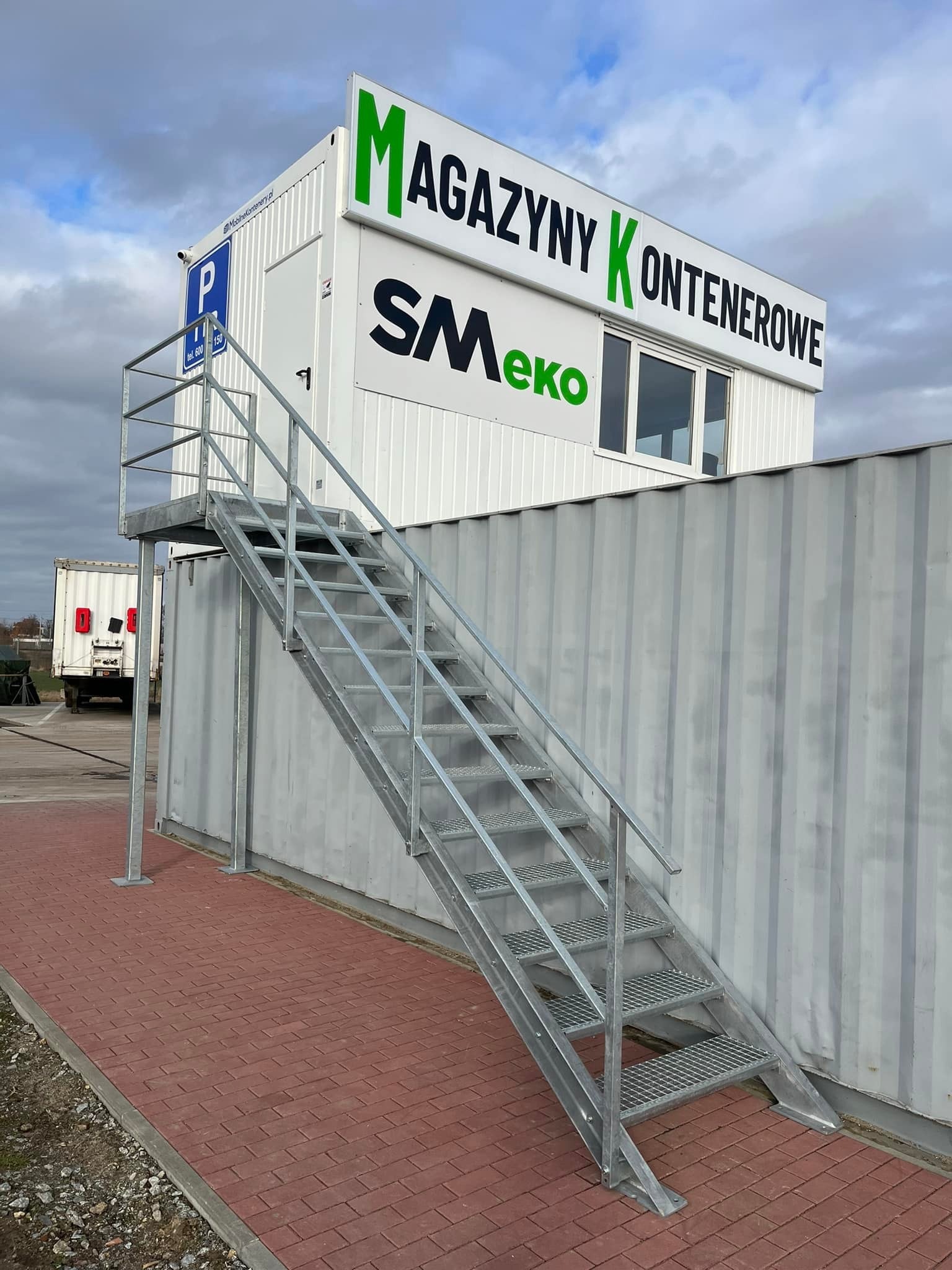 Zewnętrzne, metalowe schody z poręczą prowadzące do kontenerowego biura we Wrocławiu. Konstrukcja ocynkowana, z kratowymi stopniami. Widoczne oznakowanie magazynów.