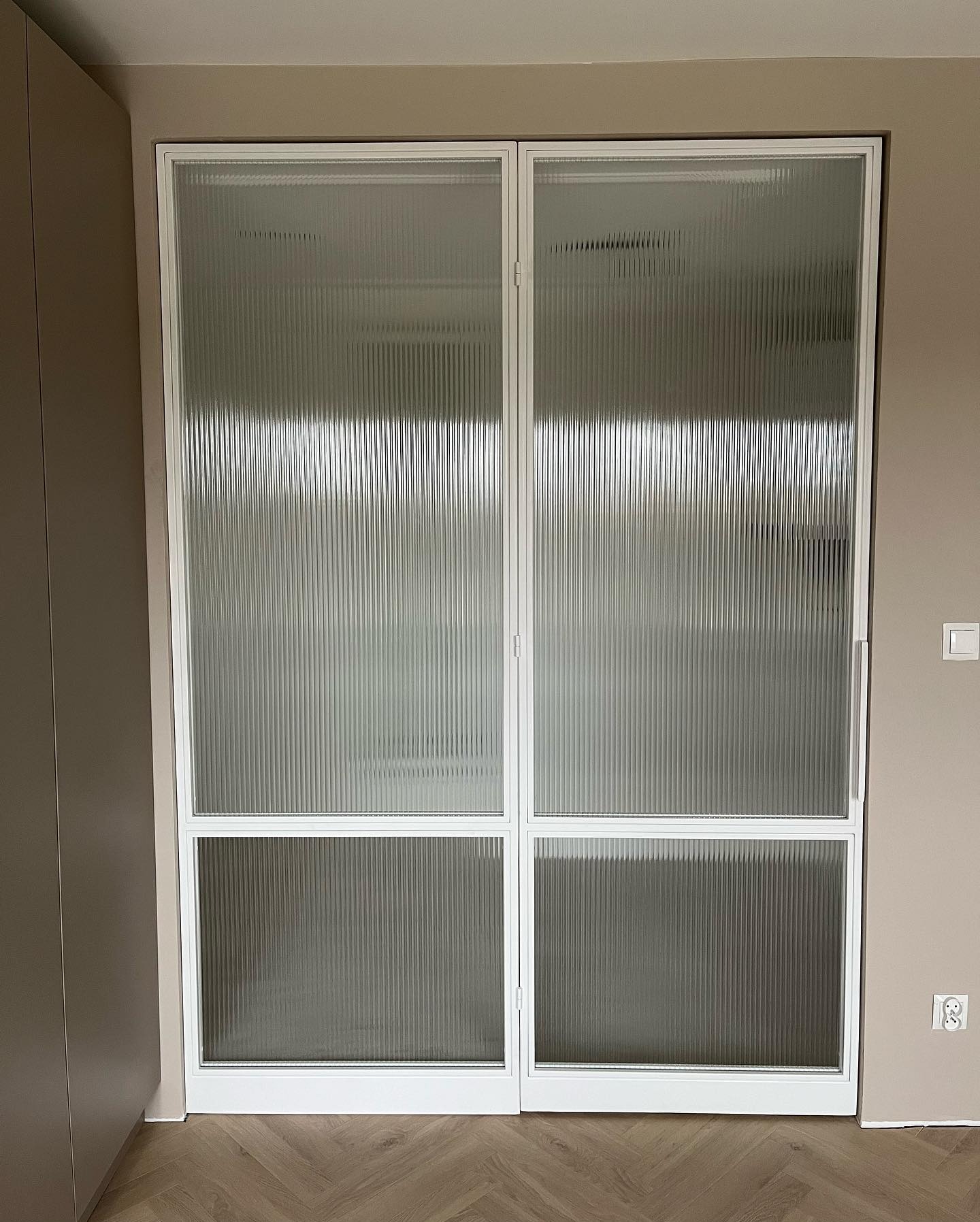 Nowoczesne, przesuwne drzwi z ryflowanego szkła w białej ramie, widoczne na tle parkietu w jodełkę i ściany w odcieniach beżu. Minimalistyczny design.