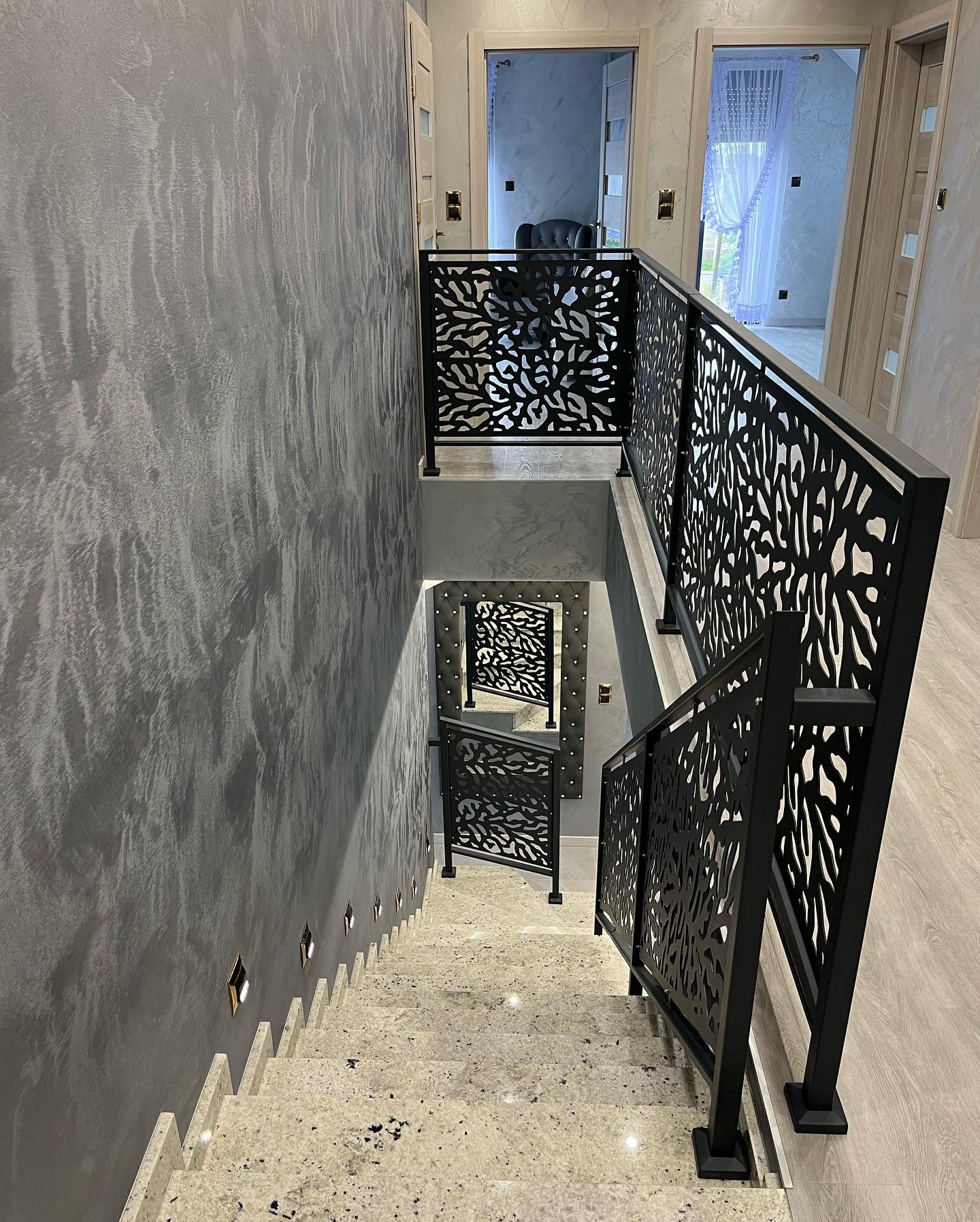 Nowoczesne schody z ażurową, czarną balustradą i kamiennymi stopniami, widok z góry. Dekoracyjna ściana z oświetleniem punktowym. Eleganckie wykończenie wnętrza.