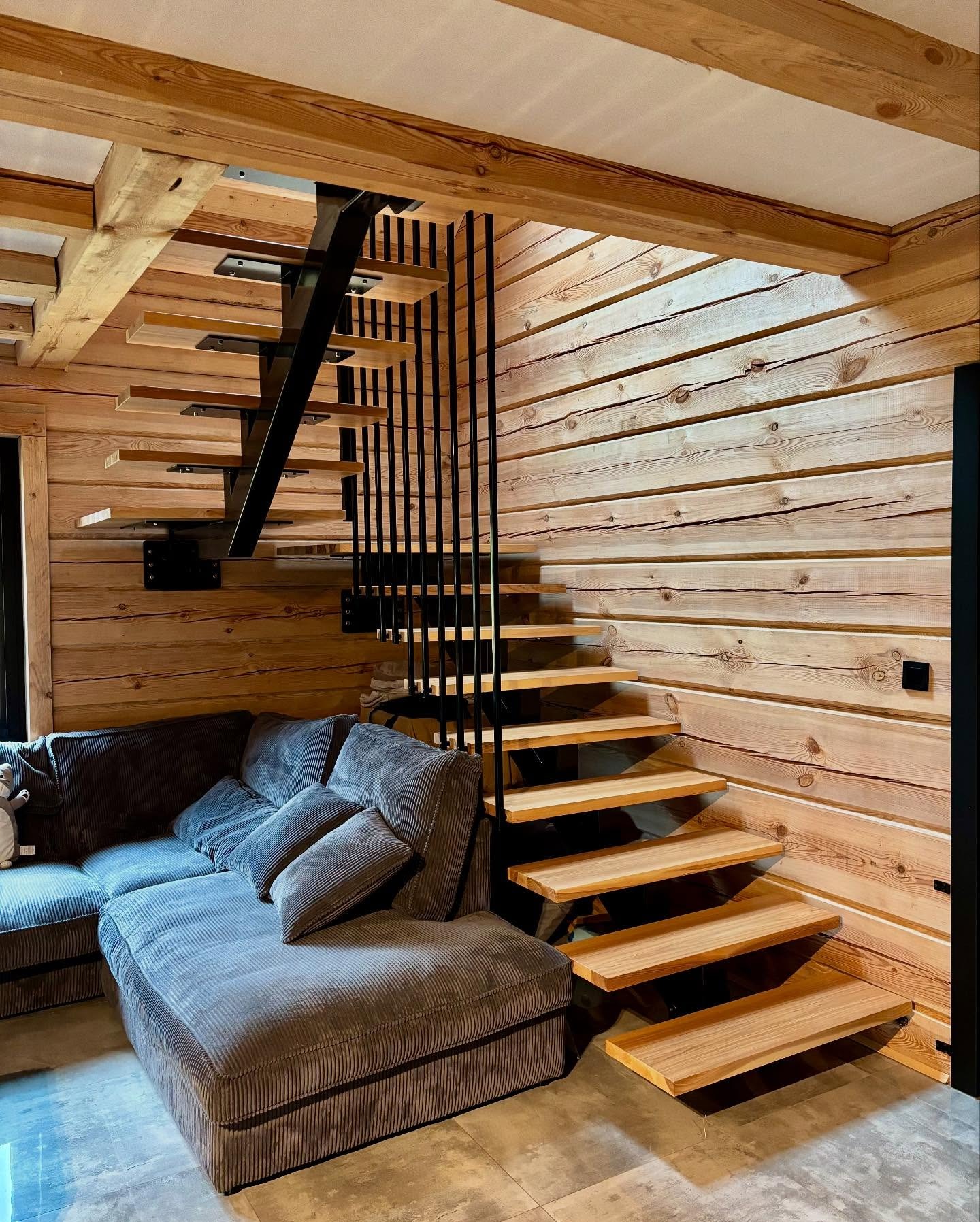 Nowoczesne schody metalowe z drewnianymi stopniami w jasnym wnętrzu z boazerią. Minimalistyczna konstrukcja i ciepłe oświetlenie tworzą przytulną atmosferę.