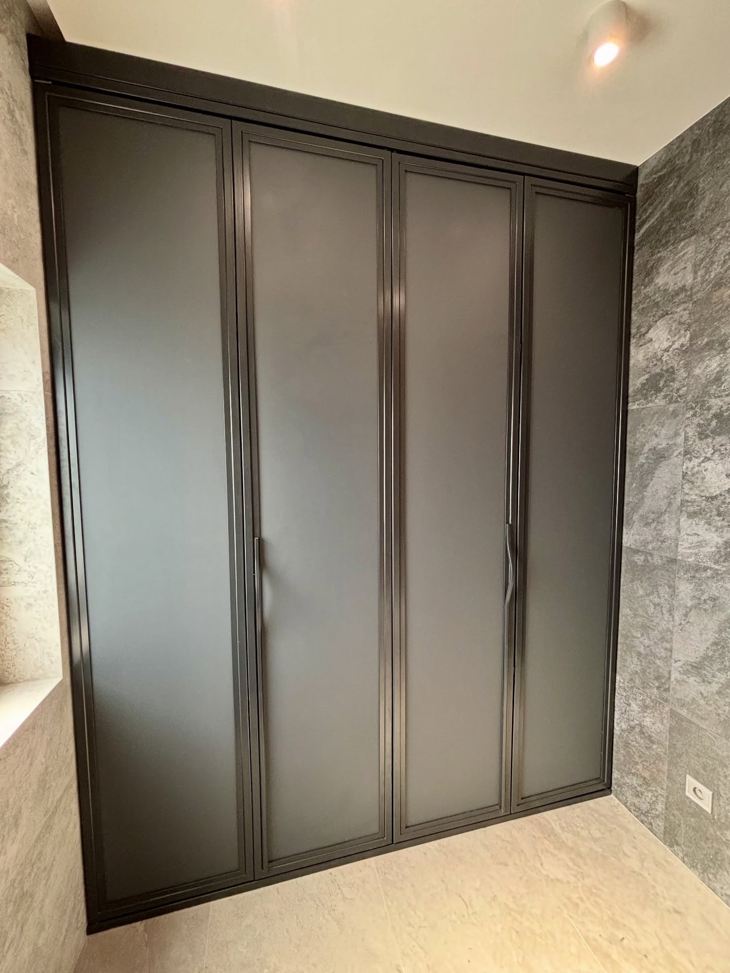 Nowoczesna, grafitowa zabudowa szafy wnękowej z metalowymi ramami i matowymi frontami, dopasowana do wnętrza z marmurowymi płytkami na ścianach.