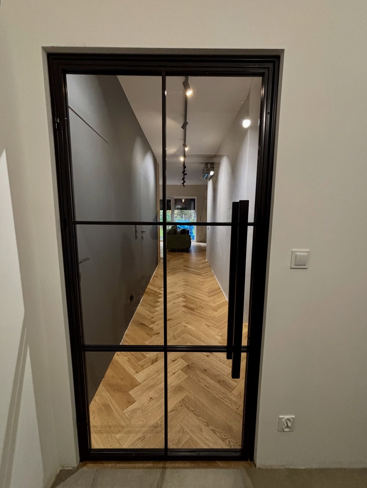 Nowoczesne, czarne, metalowe drzwi loftowe z szybami, widok na parkiet ułożony w jodełkę. Minimalistyczne wnętrze z szarą ścianą i widocznym oświetleniem.