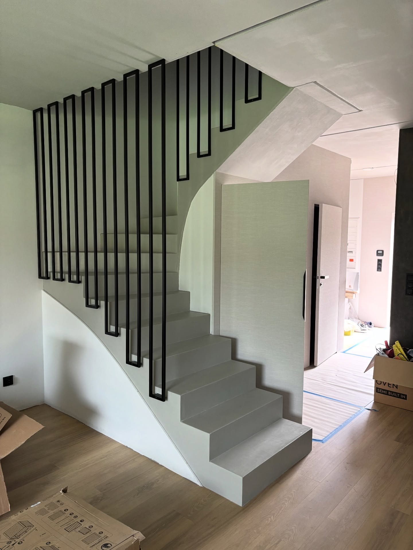 Nowoczesne schody wewnętrzne z szarą konstrukcją i czarną, minimalistyczną balustradą. Widoczne elementy wykończenia wnętrza w neutralnych barwach.