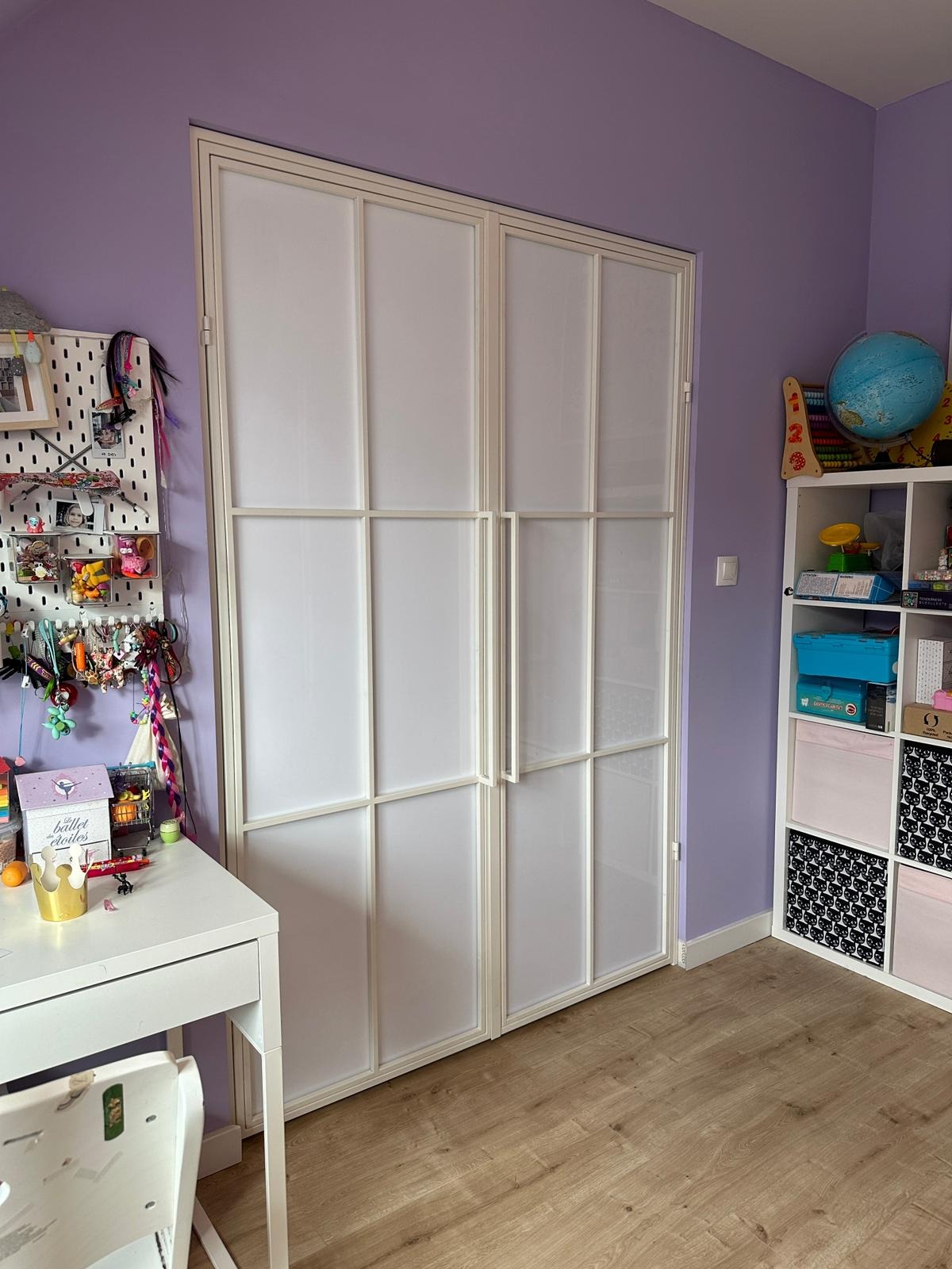 Białe, dwuskrzydłowe drzwi z metalową ramą i mlecznym wypełnieniem w pokoju dziecięcym z fioletowymi ścianami i drewnianą podłogą.