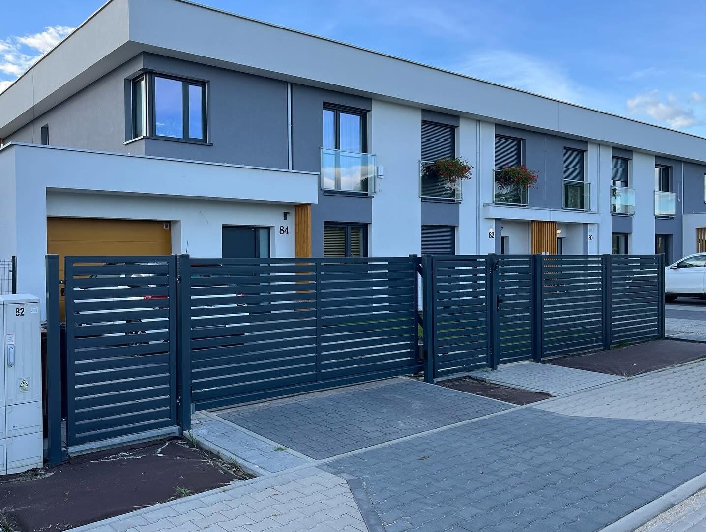 Nowoczesne, poziome ogrodzenie metalowe w kolorze grafitowym przed szeregową zabudową. Widoczne skrzydło bramy i furtka. Kostka brukowa na podjeździe.