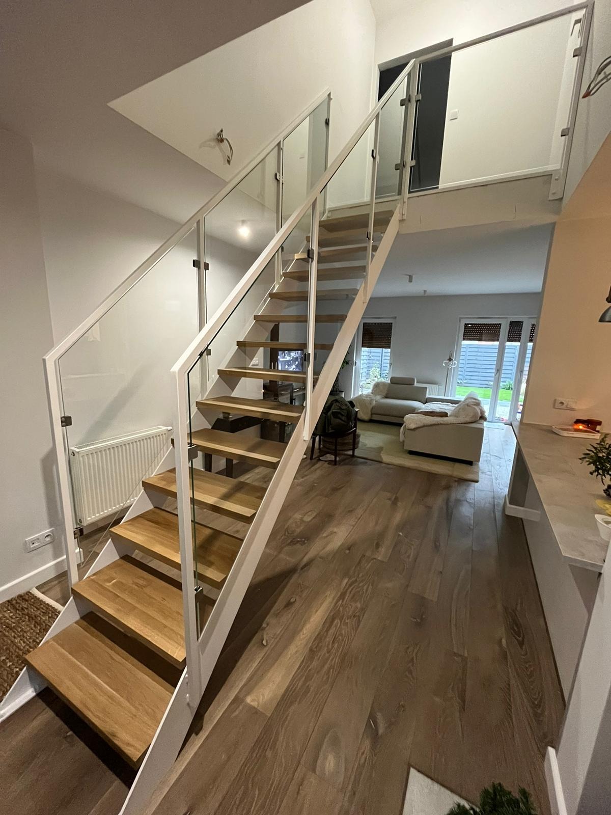 Nowoczesne schody z drewnianymi stopniami i białymi metalowymi elementami oraz szklanymi balustradami w jasnym wnętrzu domu.