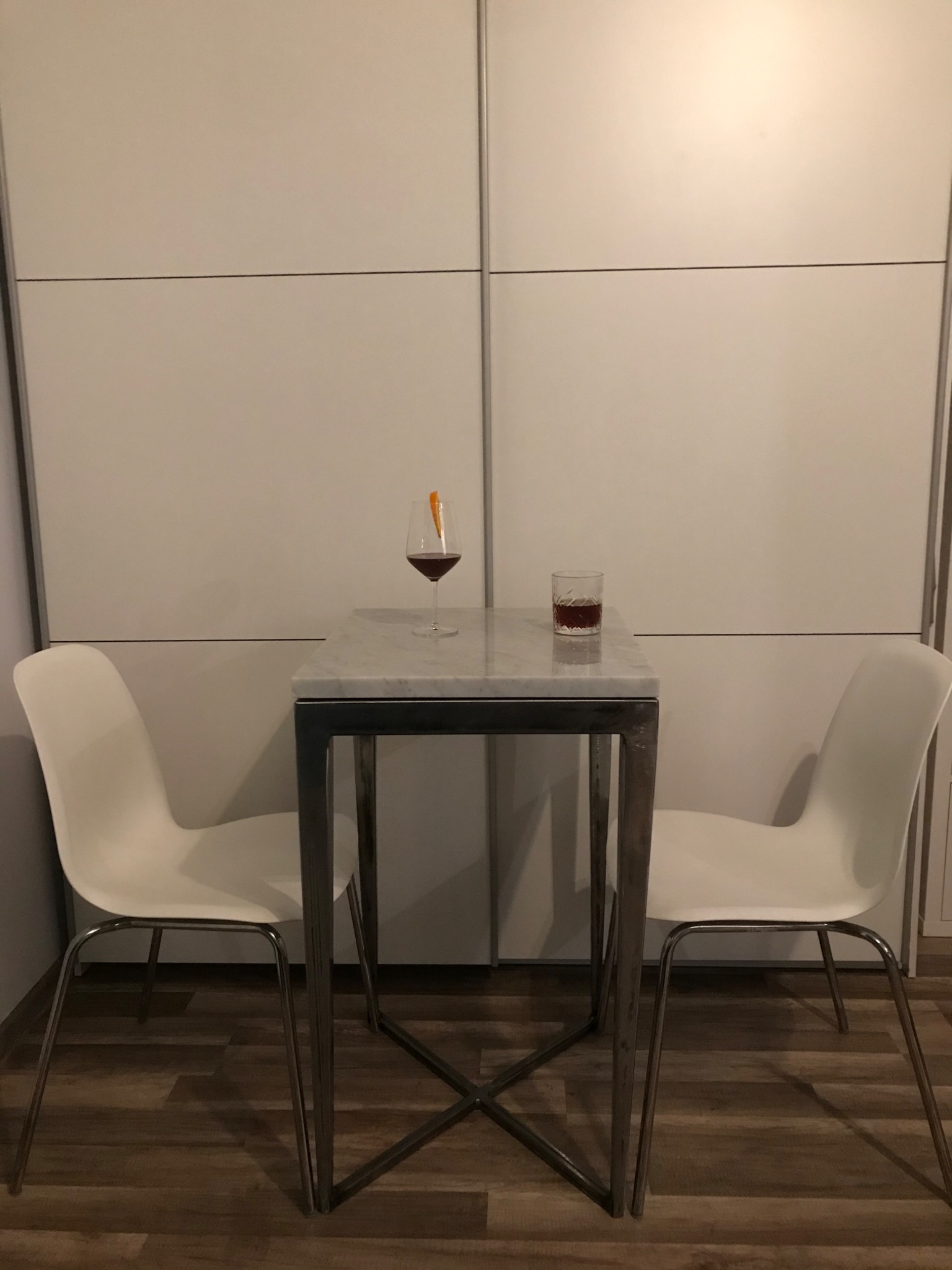 Stół barowy z marmurowym blatem i metalową konstrukcją, otoczony dwoma białymi krzesłami. Na blacie stoją dwa drinki: jeden w kieliszku z dekoracją z pomarańczy, drugi w szklance typu old fashioned...