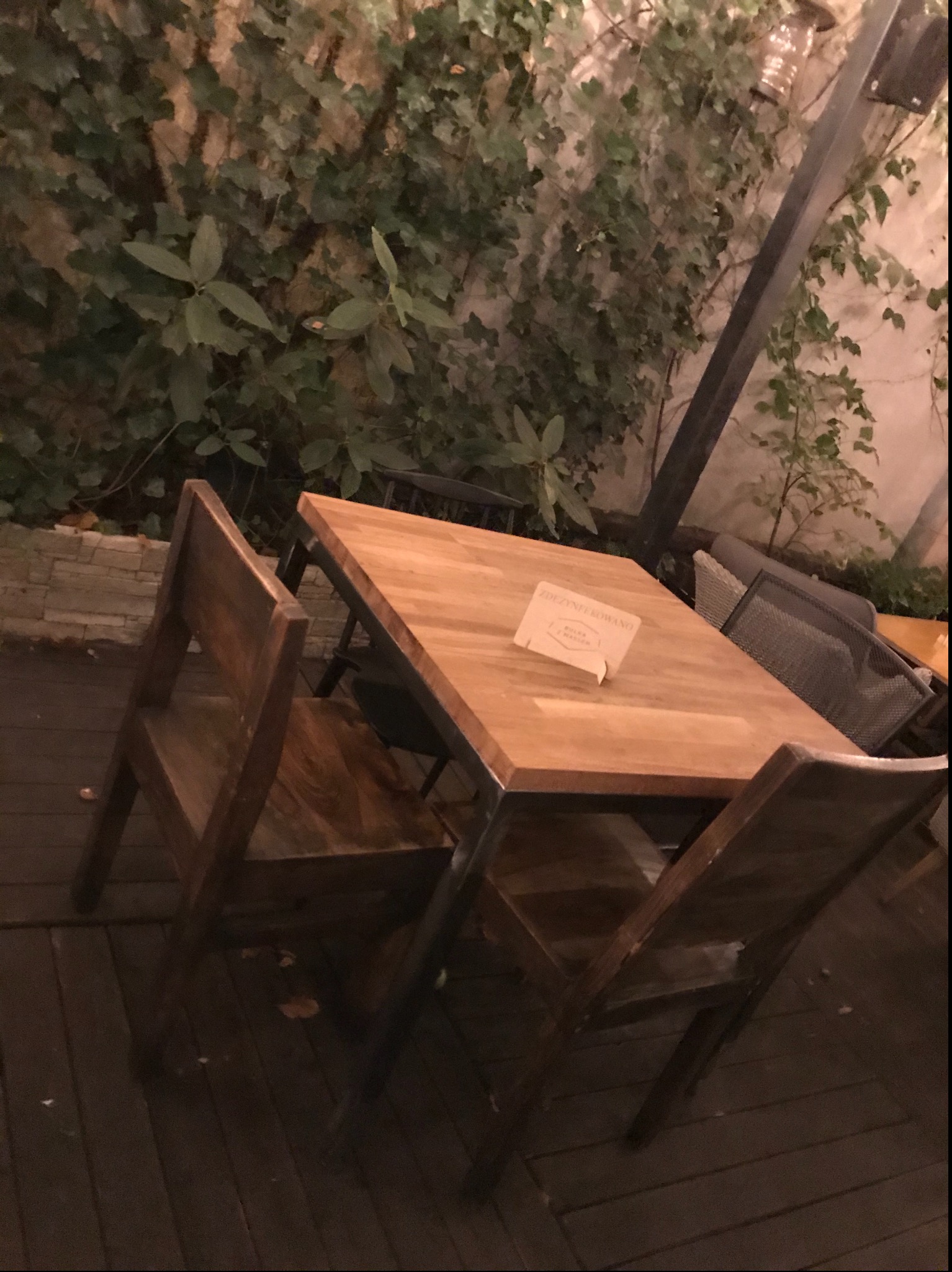 Drewniany stół i krzesła z metalowymi nogami na drewnianym tarasie, w tle ściana porośnięta bluszczem i inne meble ogrodowe.