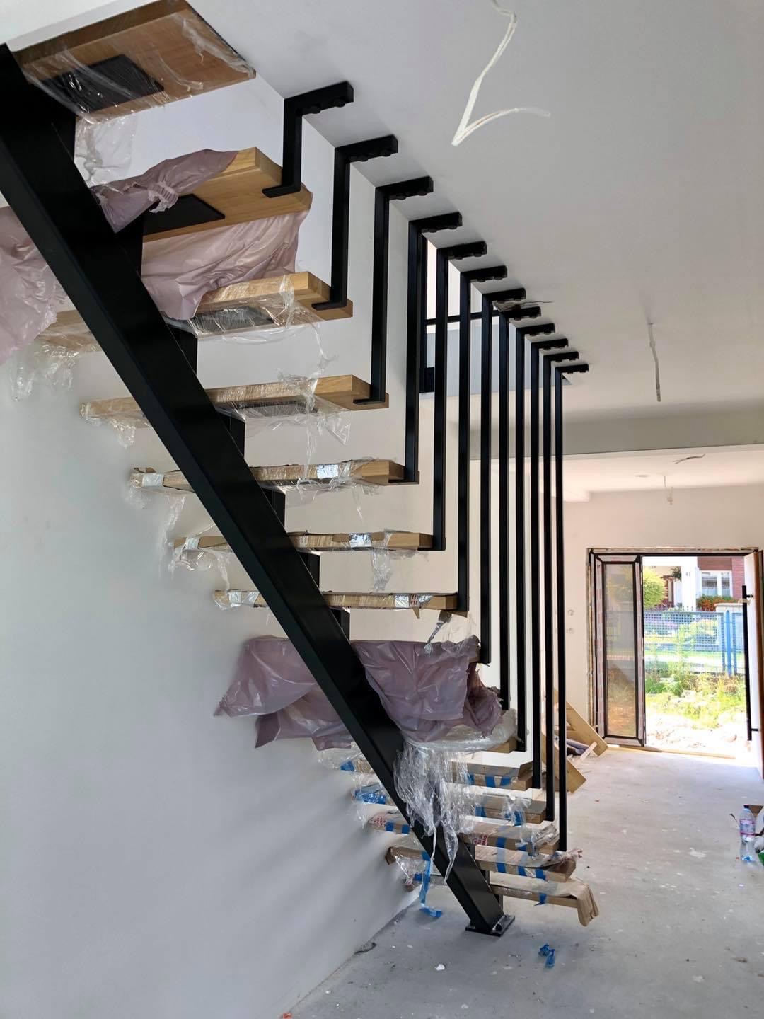 Metalowe schody z drewnianymi stopniami w trakcie montażu, zabezpieczone folią, z czarną, ażurową balustradą i widocznym fragmentem okna balkonowego w tle.