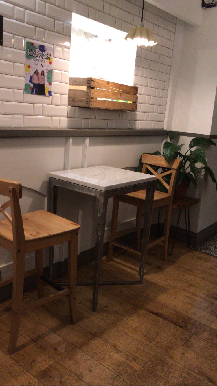Stół z marmurowym blatem i metalową ramą oraz drewniane krzesła w lokalu we Wrocławiu, z widoczną ścianą z białych płytek i drewnianą skrzynką jako elementem dekoracyjnym.