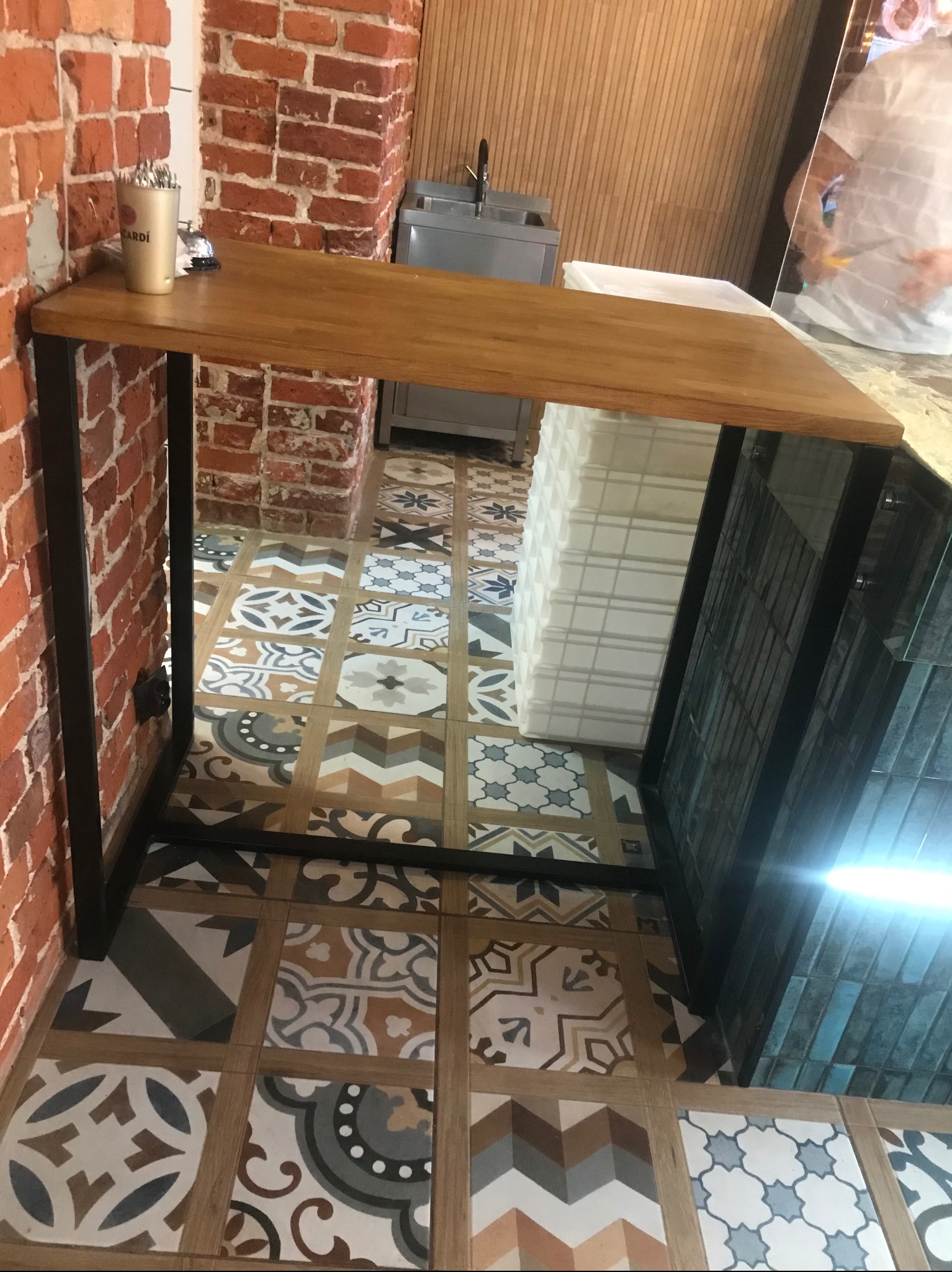 Blat drewniany na metalowych nogach, ustawiony przy ceglanej ścianie i wzorzystej podłodze, z widocznym pojemnikiem na sztućce i fragmentem zaplecza gastronomicznego w tle.