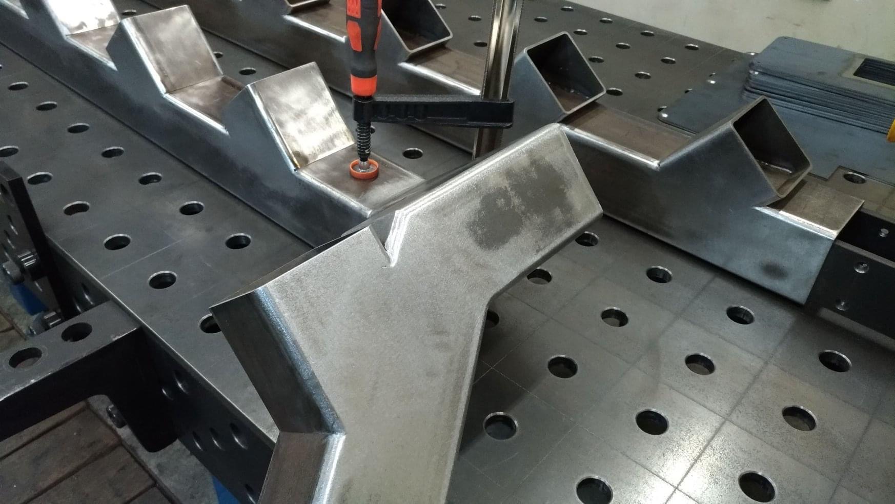 Metalowe elementy schodowe spawane i mocowane ściskiem stolarskim na perforowanym stole spawalniczym.