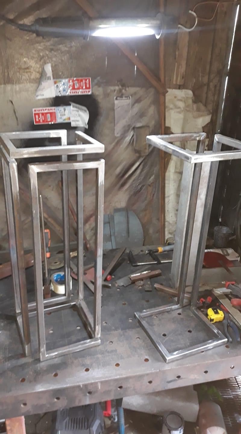 Dwie metalowe konstrukcje w kształcie prostopadłościanów z profili kwadratowych, prawdopodobnie ramy meblowe, stoją na metalowym stole warsztatowym w warsztacie, widoczne narzędzia i akcesoria.