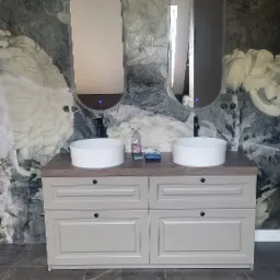 Łazienka z dwoma umywalkami nablatowymi, szafką w stylu retro z szufladami i dwoma owalnymi lustrami z podświetleniem LED na tle ściany z tapetą w kwiaty.