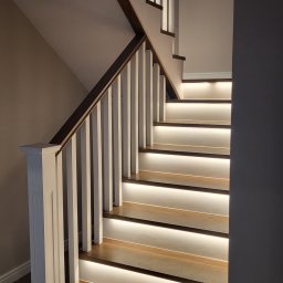 MEBLE Z PASJĄ Emilia Obłąk - Drewniane schody z białymi balustradami i podświetleniem LED na każdym stopniu, tworzącym ciepłą, nowoczesną atmosferę.